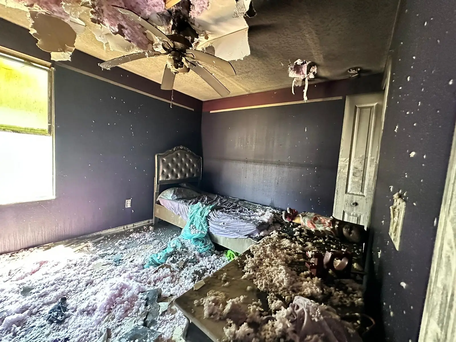 A fire-damaged bedroom with a partially collapsed ceiling, exposing insulation debris scattered across the floor and furniture. The walls are charred, and the ceiling fan is melted and warped from heat. The bed is covered in soot and debris, and personal belongings are damaged or destroyed. This image illustrates the extensive interior damage caused by a house fire, emphasizing the importance of filing a fire damage insurance claim.